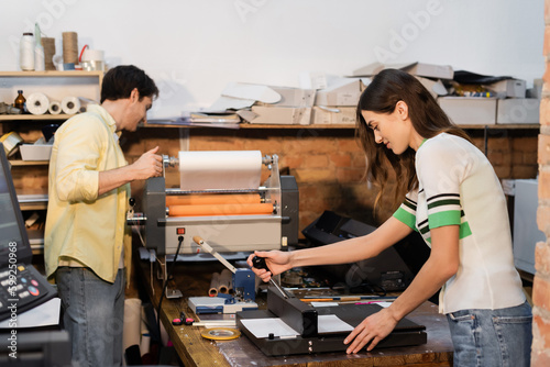 cheerful typographer using paper trimmer near colleague next to professional print plotter.