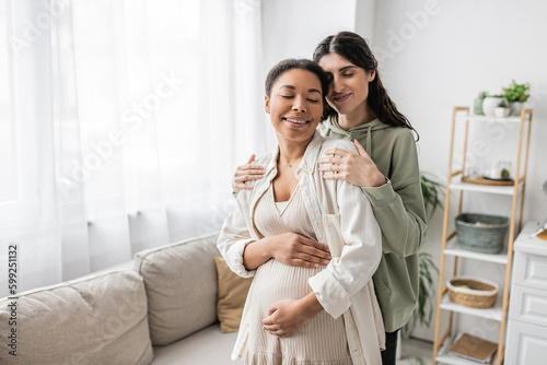 happy lesbian woman smiling while hugging pregnant multiracial wife in living room.