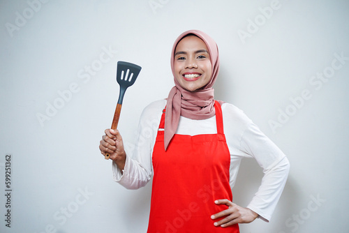 Cheerful asian muslim woman in red apron holding a spatula. Cooking ware. Cooking concept.