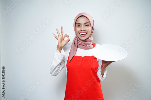 Cheerful asian muslim woman in red apron holding empty plate. Food Advertisement. Cooking concept. photo