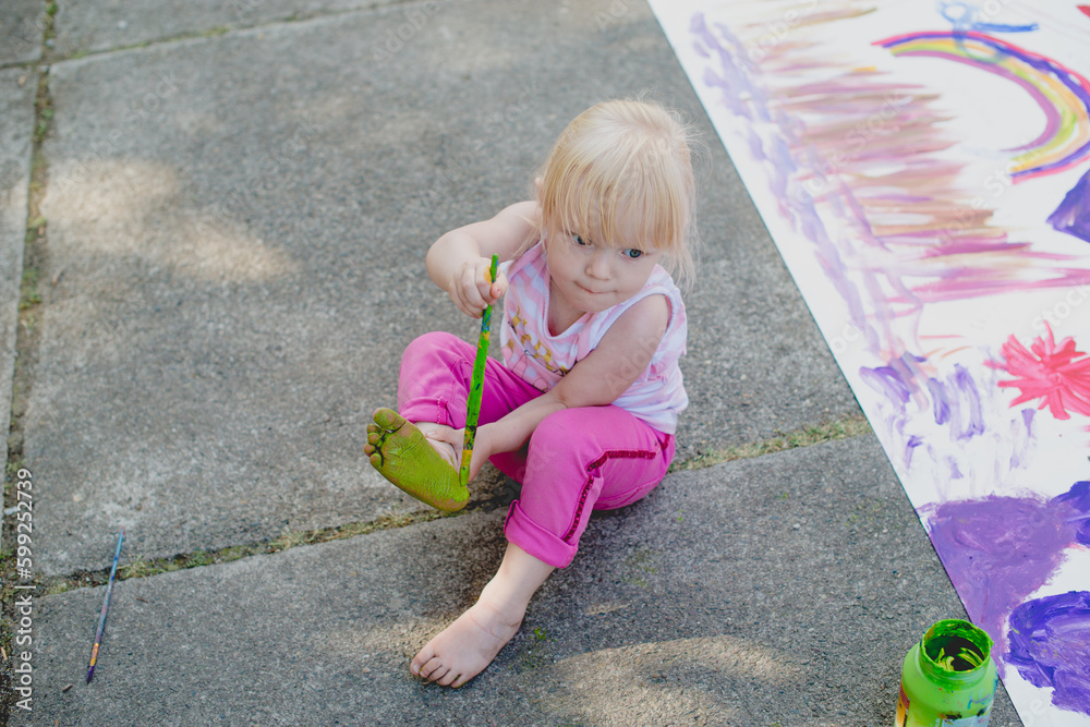 Naklejka premium A cheerful cute girl draws with green paints on her leg, the foot of her body. Toddler girl plays on the street. Real sincere moments from the life of childhood. The young artist paints on the body. F