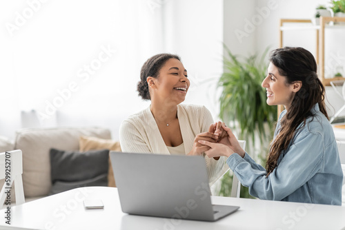 cheerful interracial lesbian couple holding hands near gadgets on table.