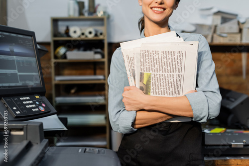 partial view of positive typographer in apron holding printed newspapers. photo