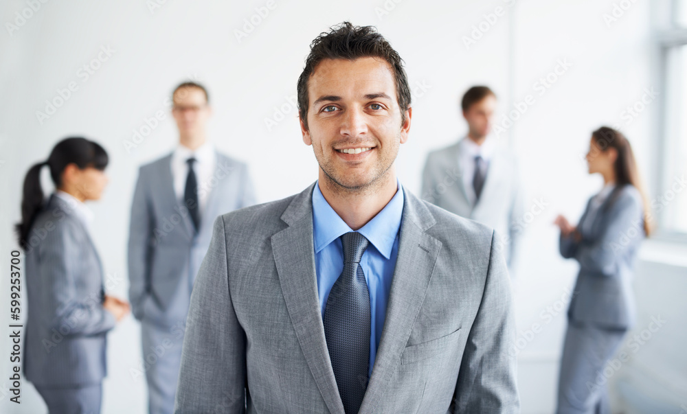 He excels at business. Portrait of a handsome young businessman smiling while his colleagues stand blurred in the background. He excels at business. Portrait of a handsome young businessman smiling while his colleagues stand blurred in the background.