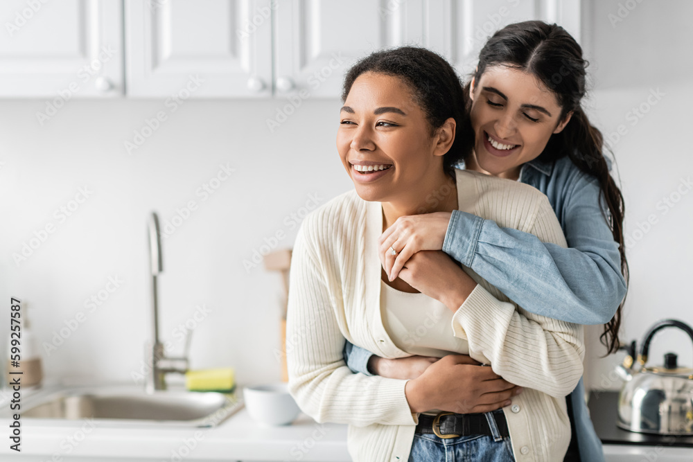 cheerful lesbian woman hugging joyful multiracial girlfriend in modern kitchen.