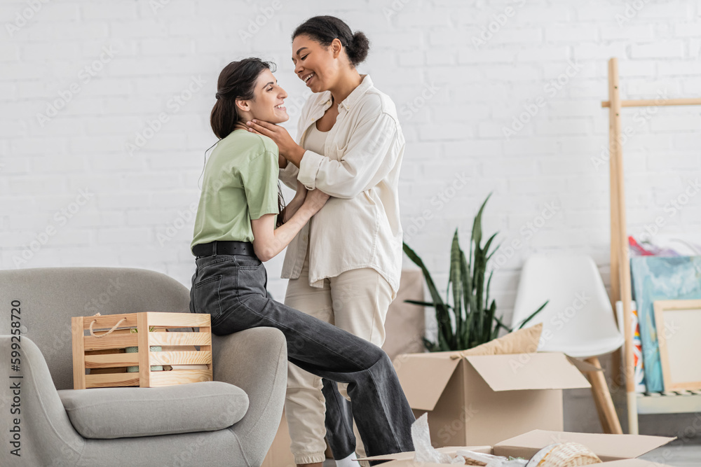 cheerful lesbian woman hugging joyful multiracial girlfriend with closed eyes in living room of new house.