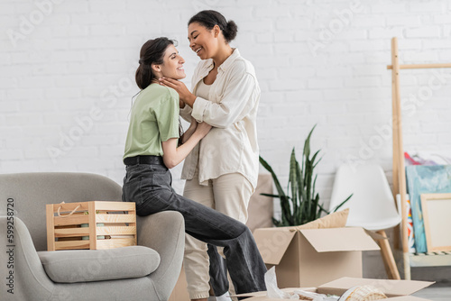 cheerful lesbian woman hugging joyful multiracial girlfriend with closed eyes in living room of new house. © LIGHTFIELD STUDIOS