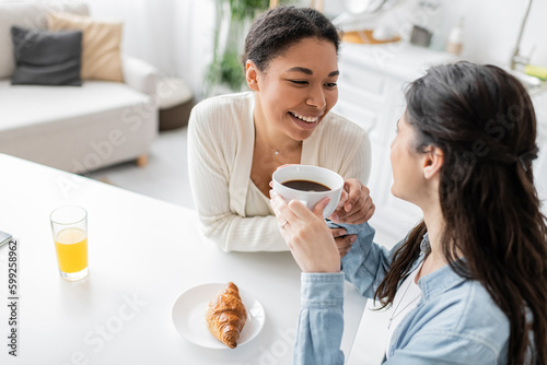 overhead view of cheerful multiracial lesbian couple smiling during breakfast in kitchen.