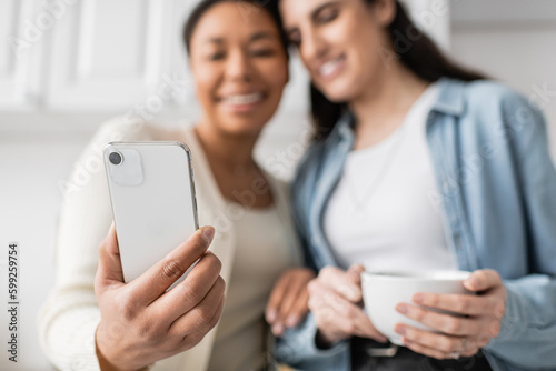 multiracial woman holding smartphone near lesbian partner with cup of coffee.