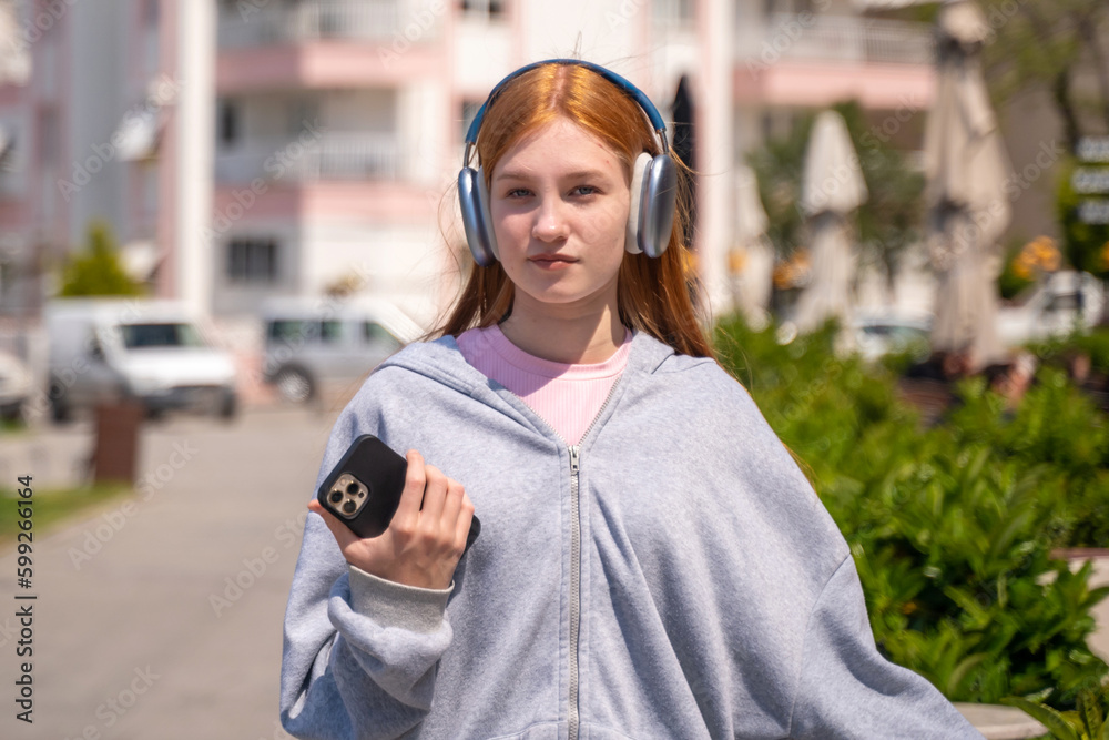 Teenage redhead girl headphones using smartphone outdoor.