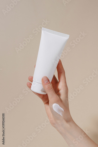 Vertical image of a female hand with a white smear holding a blank tube of moisturizing cream on a beige isolated background. Hand care, natural cosmetics and beauty concept. Image for your design