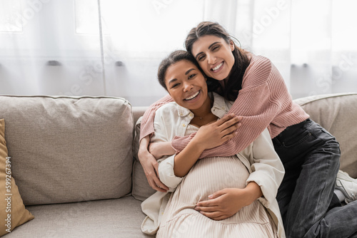 happy lesbian woman hugging pregnant multiracial partner and sitting on couch.