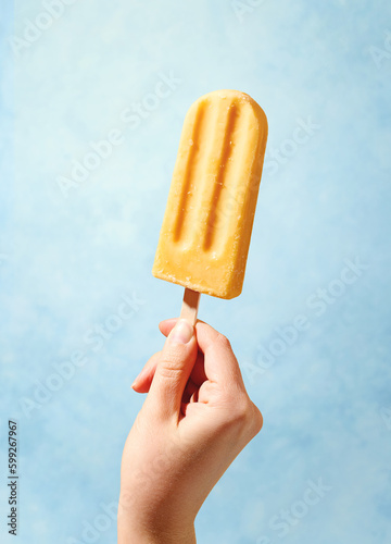 Woman hand holding yellow fruit popsicle on blue background photo