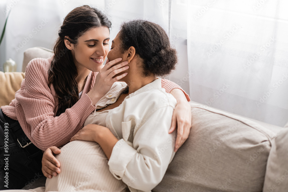 happy lesbian woman kissing pregnant multiracial partner and sitting on couch.