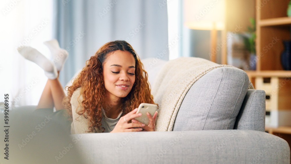Obraz premium Young woman smiling and laughing while texting on a phone at home. Cheerful female chatting to her friends on social media, browsing online and watching funny internet memes while relaxing on a sofa