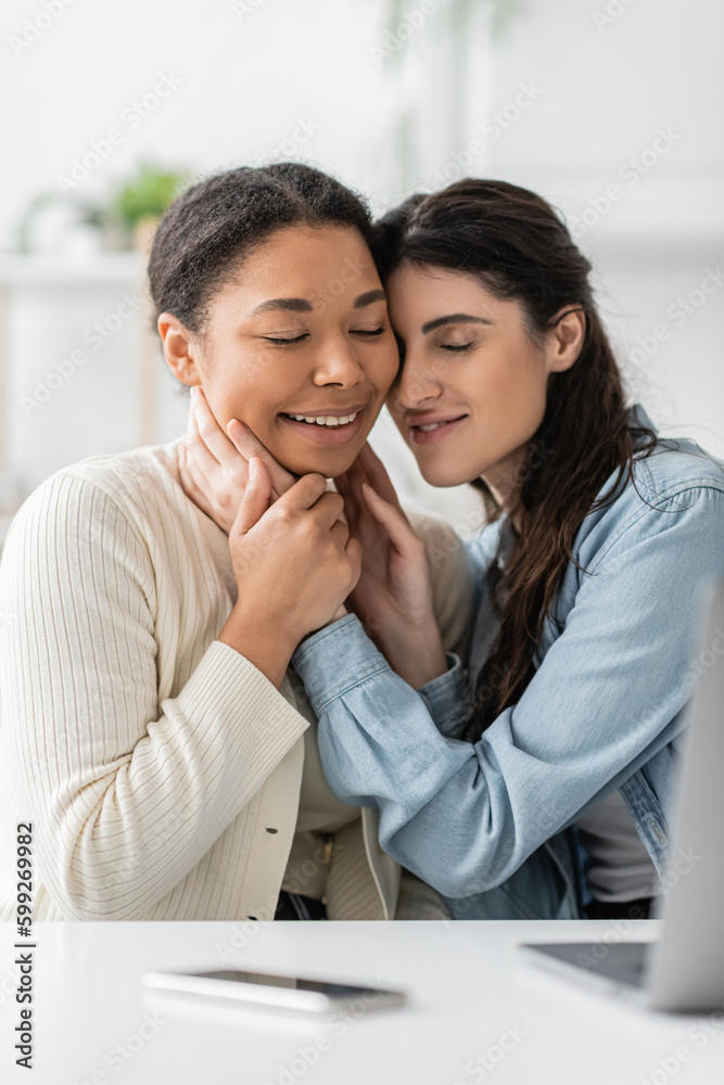 cheerful interracial lesbian couple with closed eyes hugging near devices on blurred foreground. cheerful interracial lesbian couple with closed eyes hugging near devices on blurred foreground.