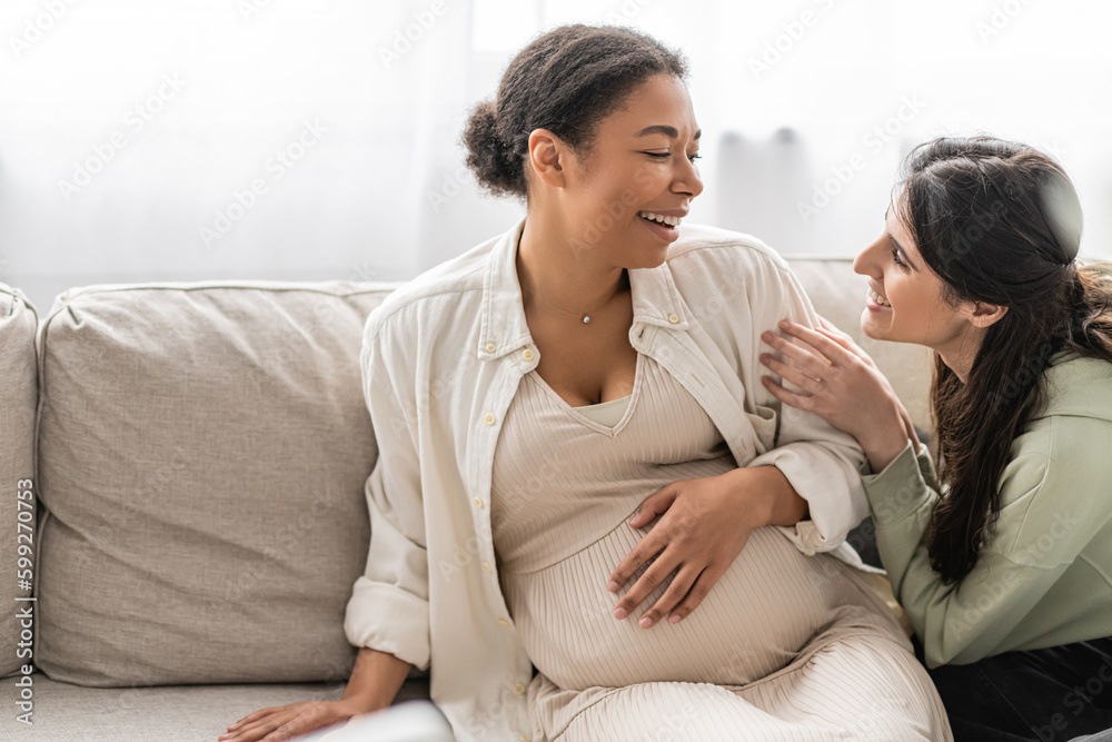 happy lesbian woman looking at cheerful pregnant multiracial wife while sitting on sofa.