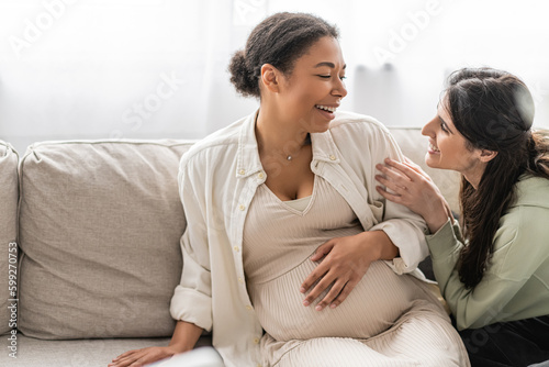 happy lesbian woman looking at cheerful pregnant multiracial wife while sitting on sofa.