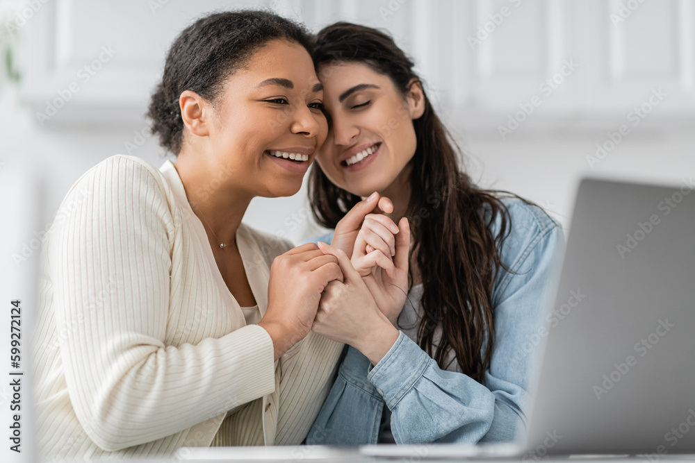pleased interracial lesbian couple holding hands near laptop. pleased interracial lesbian couple holding hands near laptop.