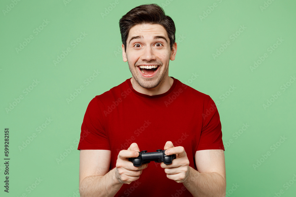 Young shocked surprised excited man wear red t-shirt casual clothes hold in hand play pc game with joystick console isolated on plain pastel light green background studio portrait. Lifestyle concept Young shocked surprised excited man wear red t-shirt casual clothes hold in hand play pc game with joystick console isolated on plain pastel light green background studio portrait. Lifestyle concept