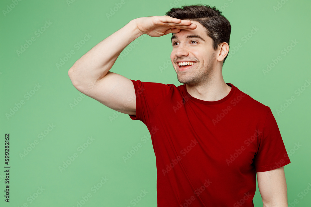 Young smiling brunet caucasian man he wears red t-shirt casual clothes hold hand at forehead look far away distance isolated on plain pastel light green background studio portrait. Lifestyle concept. Young smiling brunet caucasian man he wears red t-shirt casual clothes hold hand at forehead look far away distance isolated on plain pastel light green background studio portrait. Lifestyle concept.