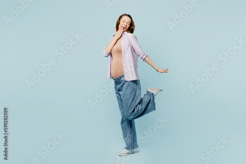 Full body side view young pregnant future mom woman with belly tummy with baby wear casual clothes looking camera hold face raise up leg isolated on plain blue background. Maternity pregnancy concept.