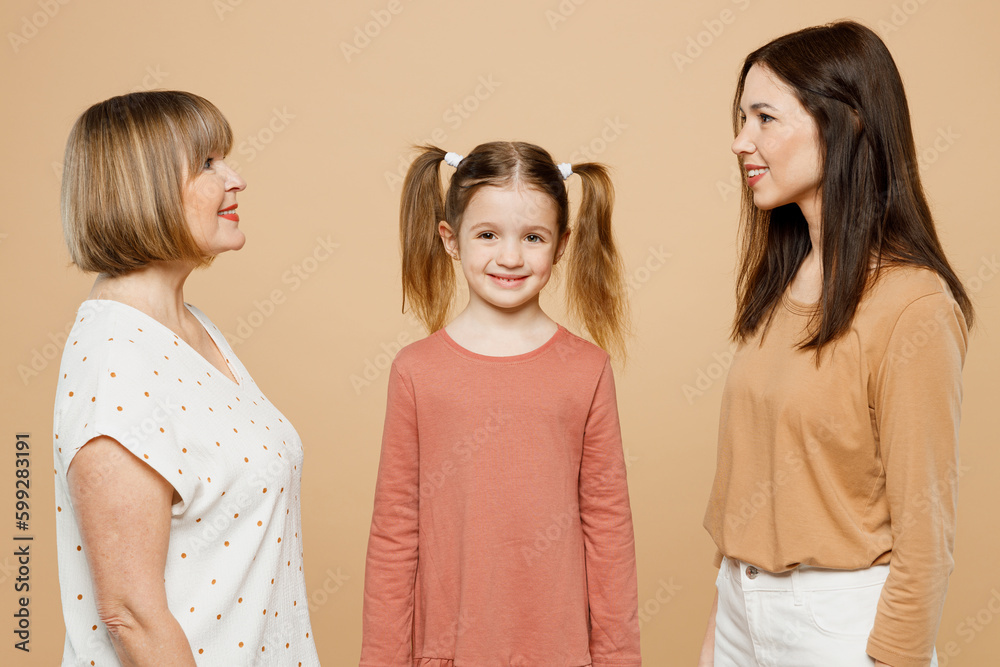 Side view happy smiling cheerful women wear casual clothes with child kid girl 6-7 years old. Granny mother daughter look to each other isolated on plain beige background. Family parent day concept. Side view happy smiling cheerful women wear casual clothes with child kid girl 6-7 years old. Granny mother daughter look to each other isolated on plain beige background. Family parent day concept.