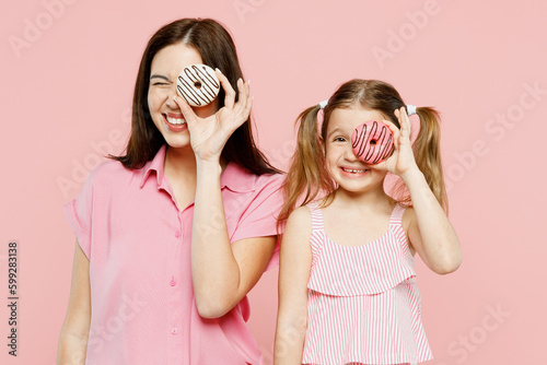 Happy cheerful woman wear casual clothes with child kid girl 6-7 years old. Mother daughter hold cover eye with donuts, look camera isolated on plain pastel pink background. Family parent day concept