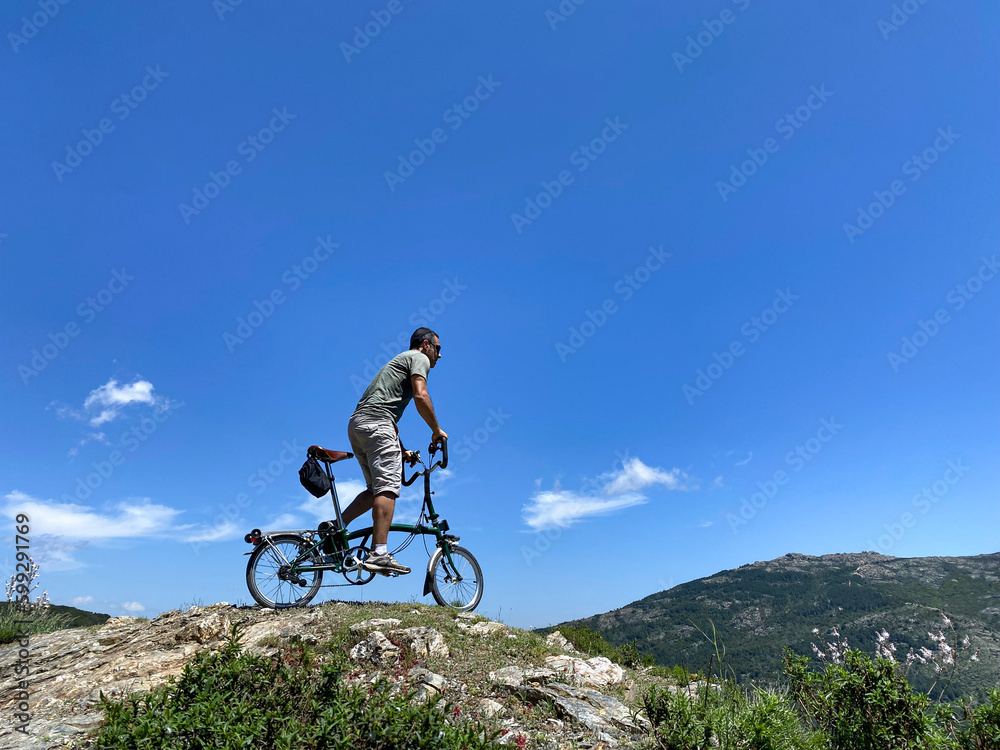 Un Uomo sulla sua bici pedala sopra una roccia, isolata su sfondo cielo azzurro