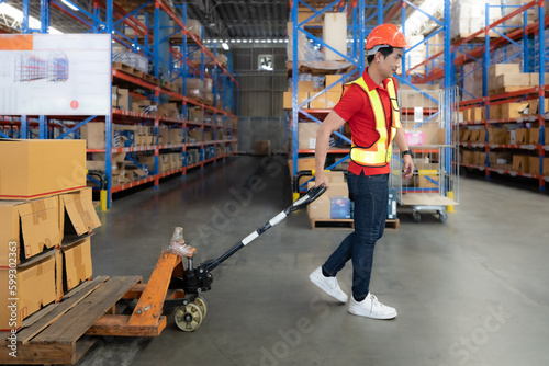 Young man pulling hand pallet truck loading package boxes stacked in shipping warehouse. Asian worker moving merchandise from storage shelf by hand lift pallet jack. Delivery goods, cargo transport. © Nassorn