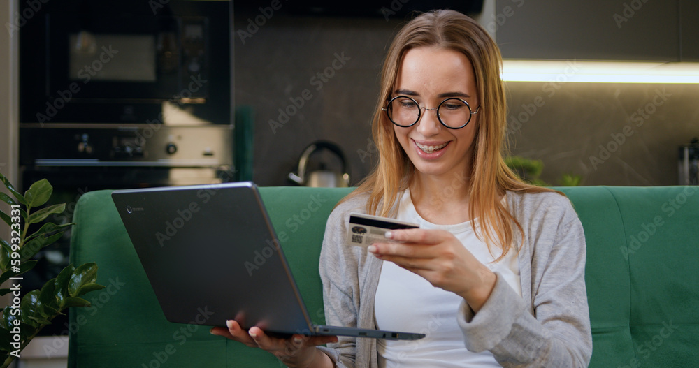 Girl doing online shopping using bank card and laptop sitting at home on sofa. Woman customer making money transaction or online internet shopping holding credit card. Girl doing online shopping using bank card and laptop sitting at home on sofa. Woman customer making money transaction or online internet shopping holding credit card.