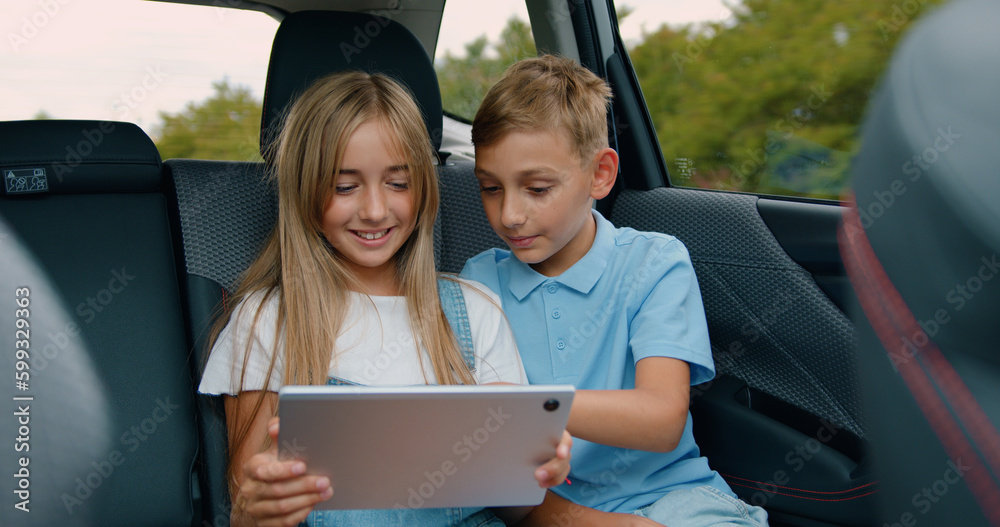 Attractive friendly smiling teen kids sitting on the rear seat in the car and playing games or watching videos on tablet pc during trip