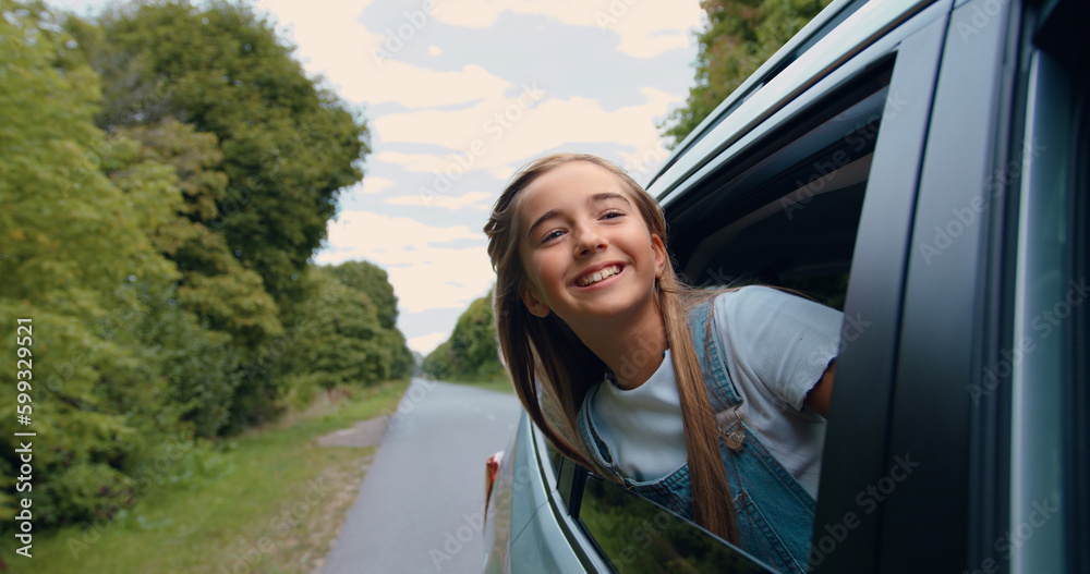 Lovely smiling happy teen girl puts her head out of the car window and enjoying surrounding nature and blowing wind through her hair during trip