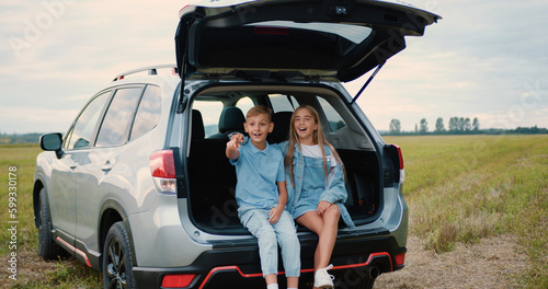 Road trip concept where good-looking positive friendly two children sitting in car trunk among green field and watching at surrounding nature © serg