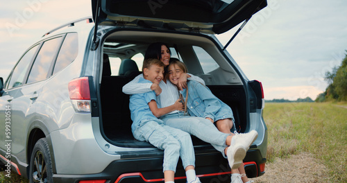 Charming loving joyful mom hugging her cheerful teen son and daughter while they together sitting in auto trunk,stopped in the green field during travel countryside,family concept