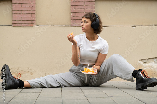woman sitting on the ground in profile, holds fruit tray and listens to music with headphones in the street
