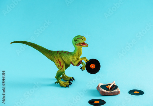 Cute green dinosaur toy holding a vinyl record and listen vinyl on vinyl record player on a blue background. © dvulikaia
