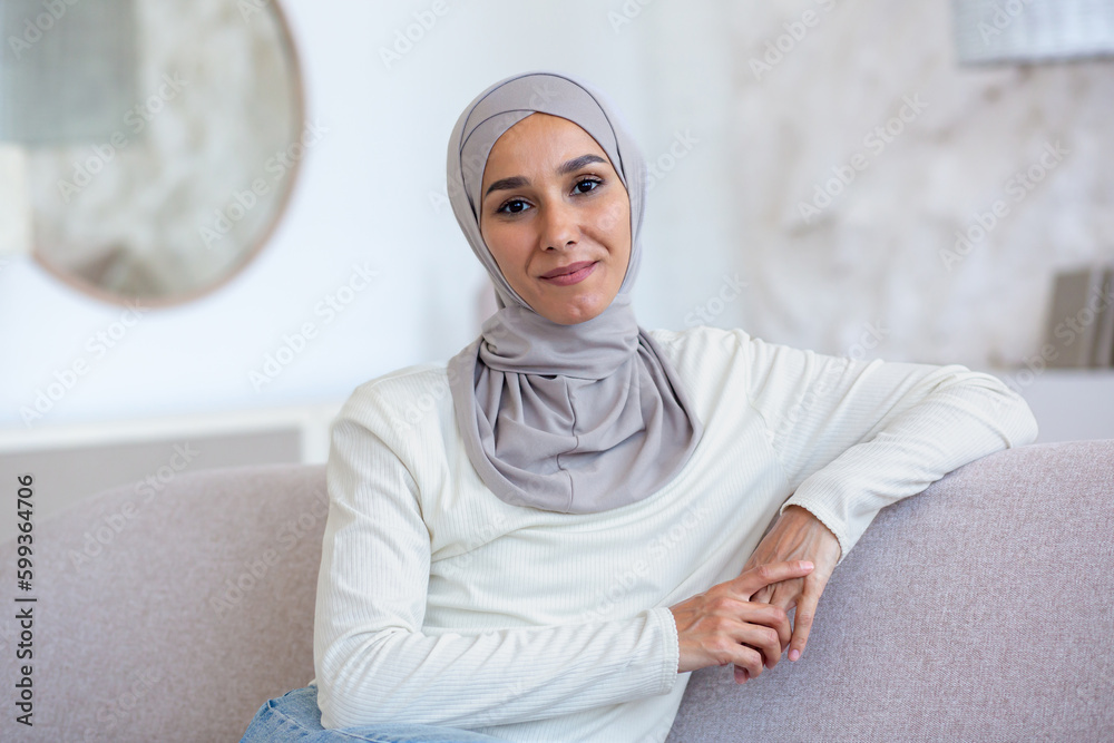Portrait of a young woman of oriental origin in a hijab sitting on the couch at home, leaning on her hand, confidently and smilingly looking at the camera.