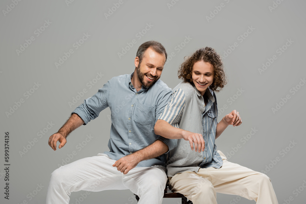 happy father and son having fun and pushing each other while sitting on one chair isolated on grey. happy father and son having fun and pushing each other while sitting on one chair isolated on grey.