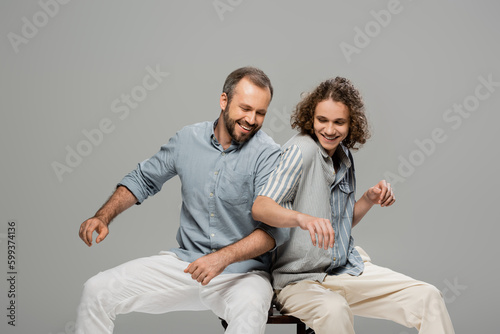 happy father and son having fun and pushing each other while sitting on one chair isolated on grey.