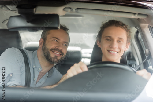 smiling teenager boy holding steering wheel while driving car next to dad.