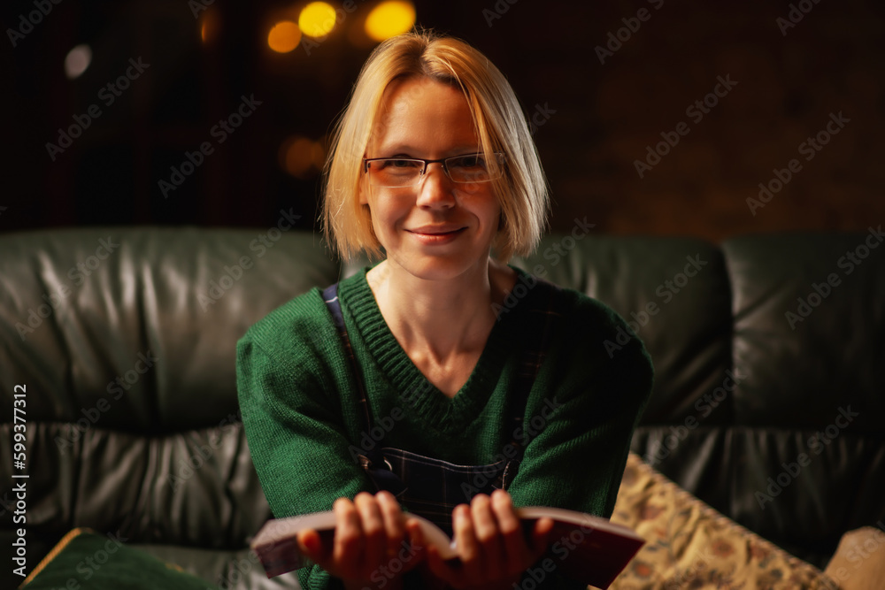 A woman in glasses smiles and looks at the camera with an open book in her hands. Middle-aged woman forty years old. Dark room, cozy atmosphere. A woman in glasses smiles and looks at the camera with an open book in her hands. Middle-aged woman forty years old. Dark room, cozy atmosphere.