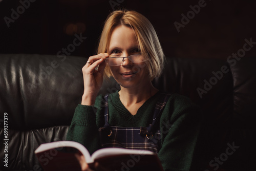 A woman in glasses smiles and looks at the camera with an open book in her hands. Middle-aged woman forty years old. Dark room, cozy atmosphere.