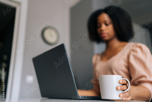 Closeup selective focus shot of black female freelancer typing on laptop looking to device screen and holding cup with coffee or tea in morning sitting at table, working from remote home office.