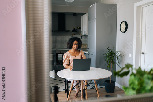Wide shot of curly African American business woman typing on laptop computer working in internet sitting barefoot at desk in kitchen. Beautiful female freelancer doing online job from home office.