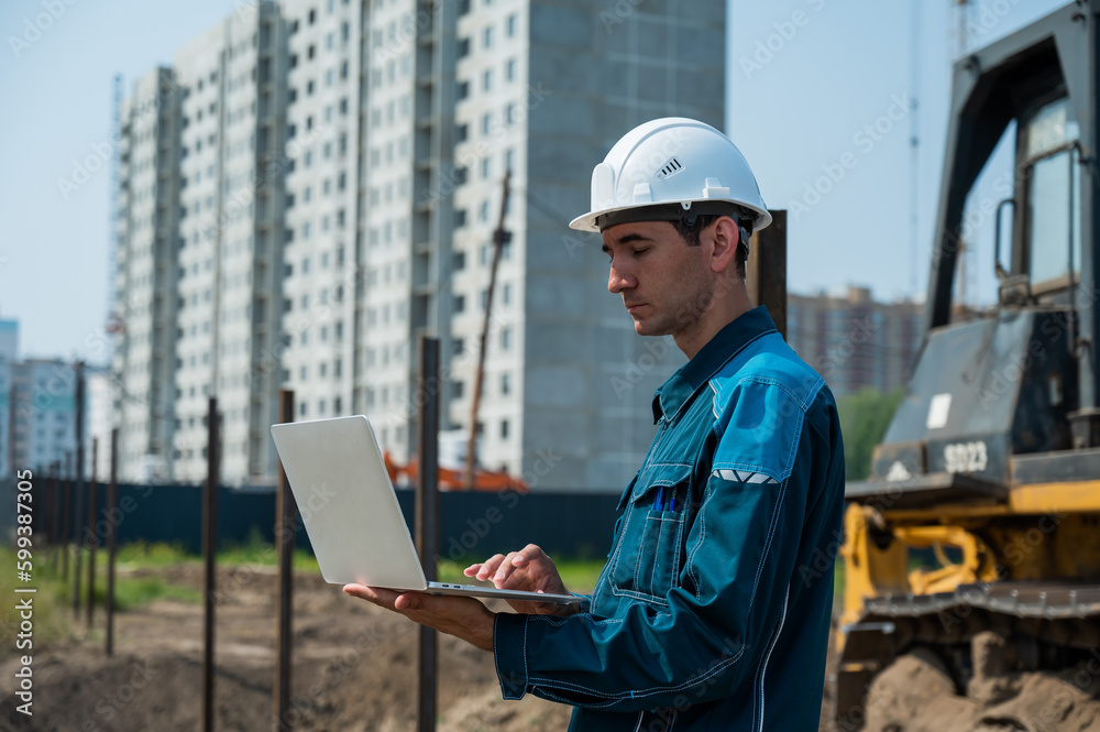 A male builder in a helmet stands against the background of a construction site with a laptop in his hands. 
