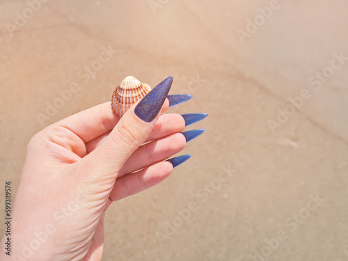 Female hand with purple stiletto nail design. Glitter purple manicure. Female hand with pink manicure hold sea shell.