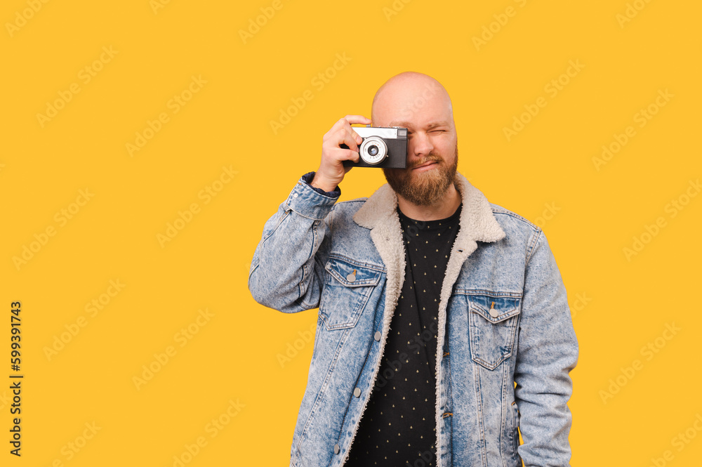 Fototapeta premium Young bald man wearing jeans jacket is taking a picture with a vintage camera over yellow background.
