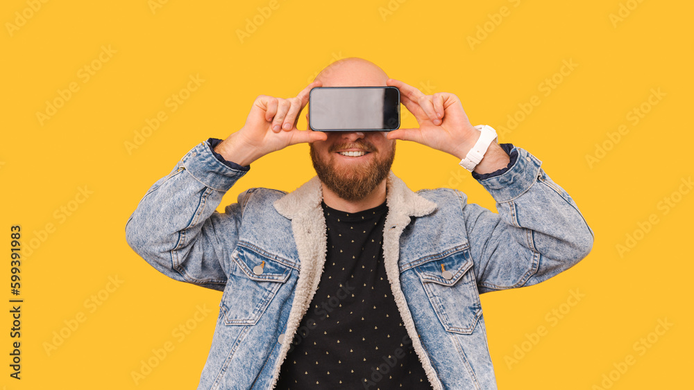 Studio portrait of a bald young men covering his eyes with a phone screen over yellow background. Studio portrait of a bald young men covering his eyes with a phone screen over yellow background.