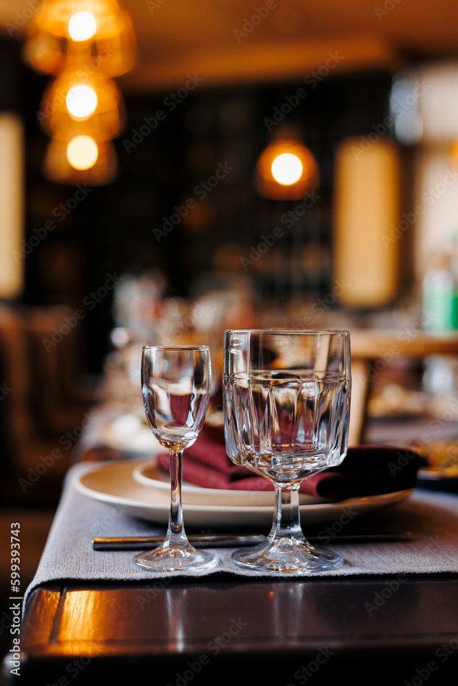 Festively table setting in a restaurant, close up two empty shiny wine glasses. Served table for wedding, birthday, anniversary party. Plates, cutlery, red napkins on wooden table. Soft electric light